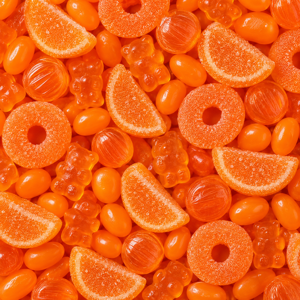 A close-up, top-down view of assorted orange candies filling the frame, including gummy bears, ring gummies, citrus slice gummies, and translucent orange jelly beans.