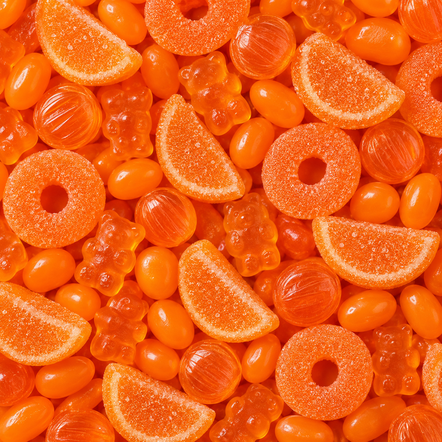 A close-up, top-down view of assorted orange candies filling the frame, including gummy bears, ring gummies, citrus slice gummies, and translucent orange jelly beans.